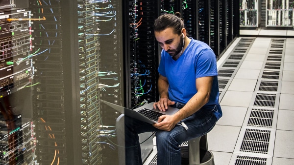 A man in a blue T-shirt and jeans sitting on a stool in a Data Center working on his laptop in networking.