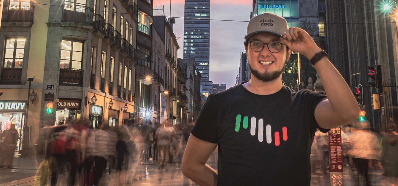 Male wearing a Cisco Hat and shirt holding left hand to his hat on a street in Mexico City